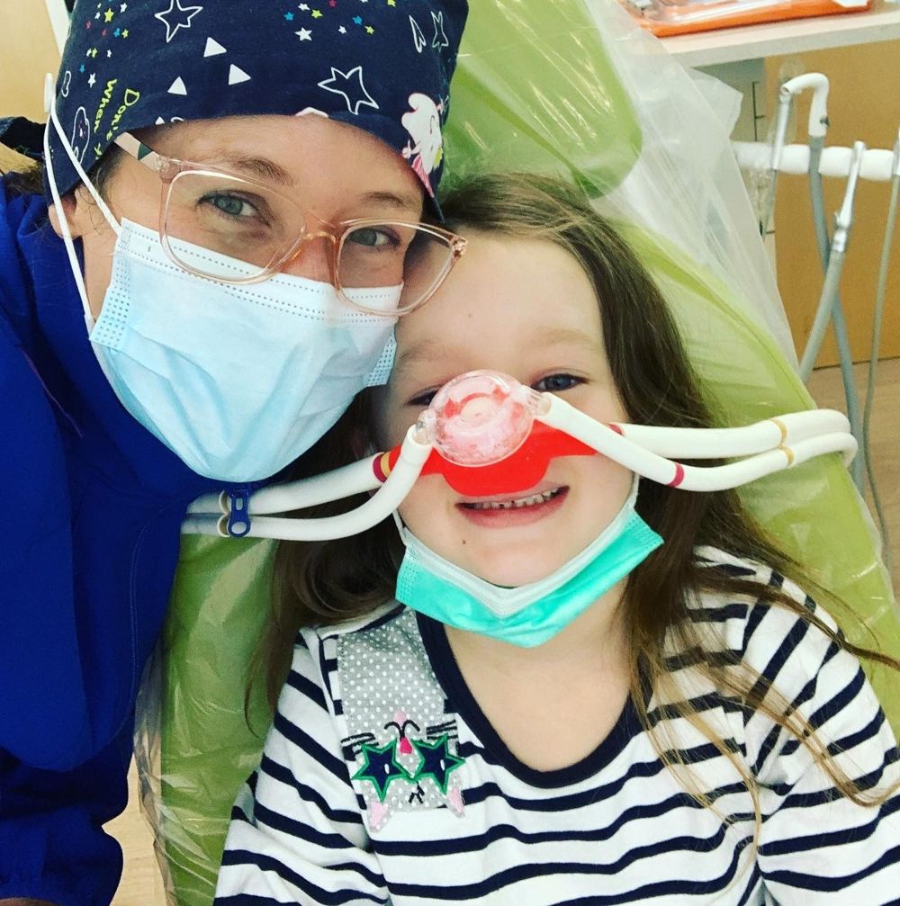 A smiling girl in the dental chair wearing a laughing gas nosepiece, cheek to cheek with her dentist
